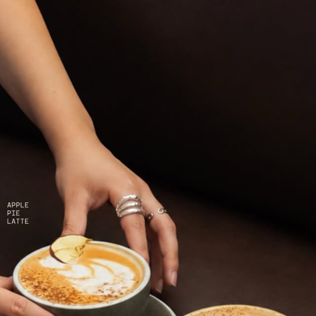 Mano sirviendo un Apple Pie Latte con arte latte y rodaja de manzana deshidratada sobre fondo café oscuro